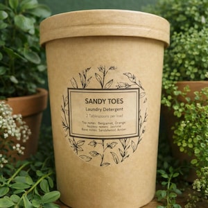 May include: A cylindrical, light brown container of "SANDY TOES Laundry Detergent" with a lid. The label includes the text "2 Tablespoons per load" and scent notes. The container is surrounded by green plants in pots.