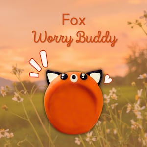 Può includere: Un Worry Buddy a forma di volpe arancione con orecchie e occhi neri e bianchi. Il testo "Fox Worry Buddy" è visualizzato sopra la volpe. Lo sfondo è un'immagine sfocata di un campo con fiori.