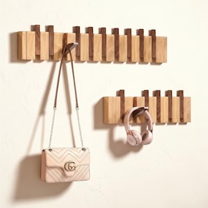 Op de afbeelding: Twee houten kapstokken met een geometrisch ontwerp. Eén kapstok houdt een lichtroze handtas met een zilveren kettingriem. De andere kapstok houdt een paar roze koptelefoons. De kapstokken zijn gemaakt van licht en donker hout.