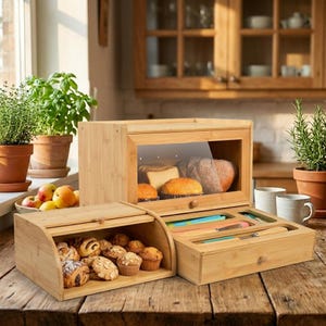 May include: Three bamboo bread boxes and a drawer with utensils on a wooden table. One box has a glass front and contains bread. Another box holds pastries. The drawer has colourful cutting boards and a knife.