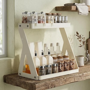 Puede incluir: Un especiero de dos niveles, de color blanco roto, con varios frascos y botellas de vidrio. La balda superior contiene pequeños frascos con especias y recipientes metálicos. La balda inferior tiene botellas de aceite y vinagre, y frascos con nueces y granos de pimienta.