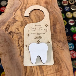 May include: Wooden door hanger with a tooth-shaped cutout and the text "Welcome Tooth Fairy Please Stop Here For: Lilly". The hanger is light brown with a white tooth and star accents.