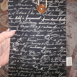 Könnte beinhalten: Schwarze Tablet-Hülle mit weißer Schrift und Holzknopfverschluss. Die Hülle hat eine beige Kordelschlaufe und ein kleines Etikett mit der Aufschrift "Handmade with care by Tonya Marie."