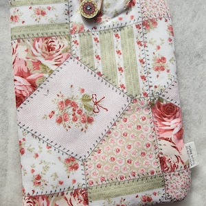 May include: A patchwork tablet sleeve with a floral rose design in pink, red, and green hues. It has a wooden button and loop closure. The design incorporates diverse fabric patterns and stitched seams. The sleeve is a charming accessory.