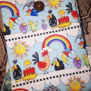 Könnte beinhalten: Ein Stoff-Buchumschlag mit einem skurrilen Design mit Cartoon-Vögeln, Regenbogen und Sonnen auf hellblauem Hintergrund. Der Umschlag wird mit einem braunen Knopf und einem Kordelverschluss gesichert. Das Design beinhaltet Vögel mit Regenschirmen.