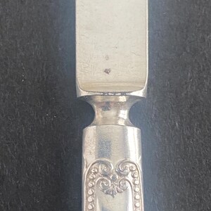 May include: Close-up of a silver butter knife. The knife has a rectangular blade and a decorative handle with an ornate design. The handle features a series of small, raised dots along the edges of the design. The knife is made of a shiny, reflective metal.