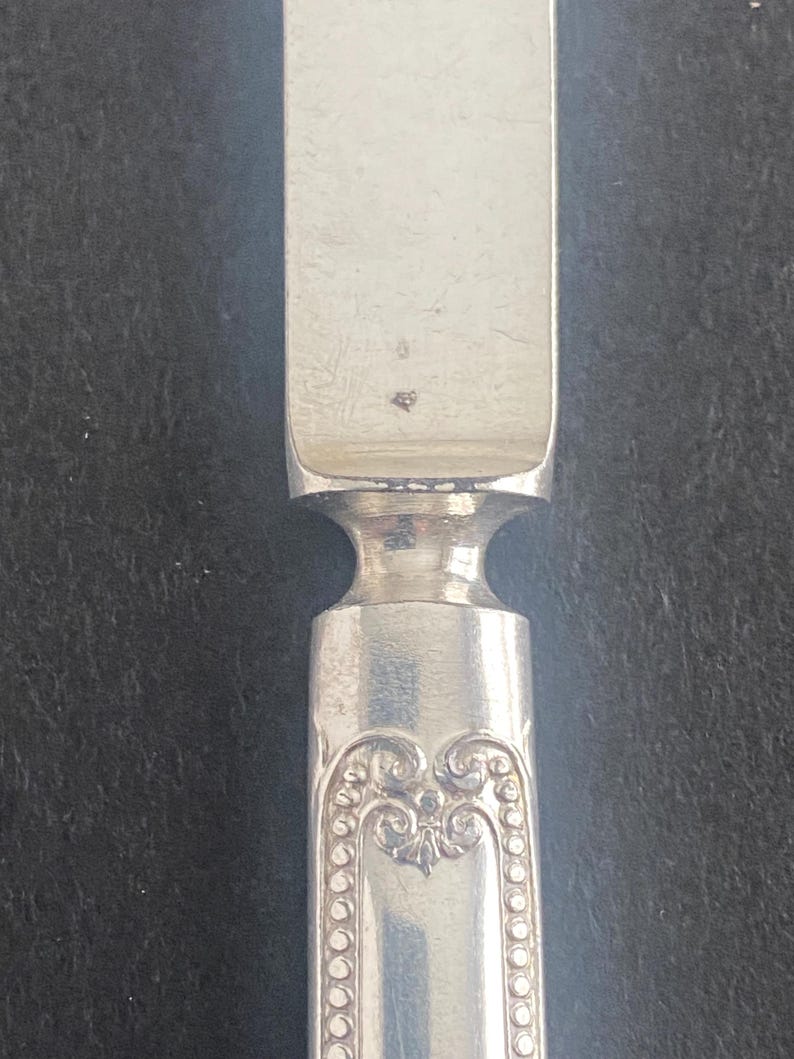 May include: Close-up of a silver butter knife. The knife has a rectangular blade and a decorative handle with an ornate design. The handle features a series of small, raised dots along the edges of the design. The knife is made of a shiny, reflective metal.