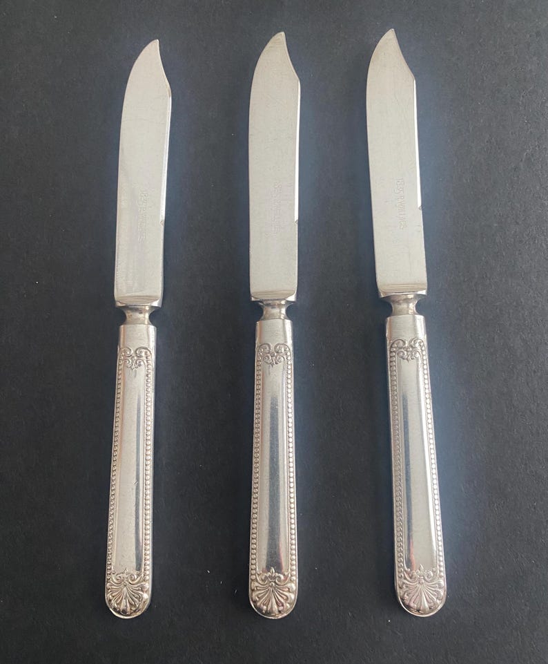 May include: Three silver butter knives with curved blades and ornate handles. The handles have a detailed, symmetrical design with a floral motif at the base. The blades are smooth and shiny, reflecting the light. The knives are arranged on a dark surface.