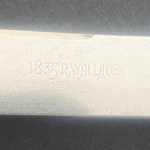 May include: Close-up of a silver metal object with the engraved text "1835 R. WALLACE". The metal surface has visible scratches and wear, indicating age. The text is in a serif font.