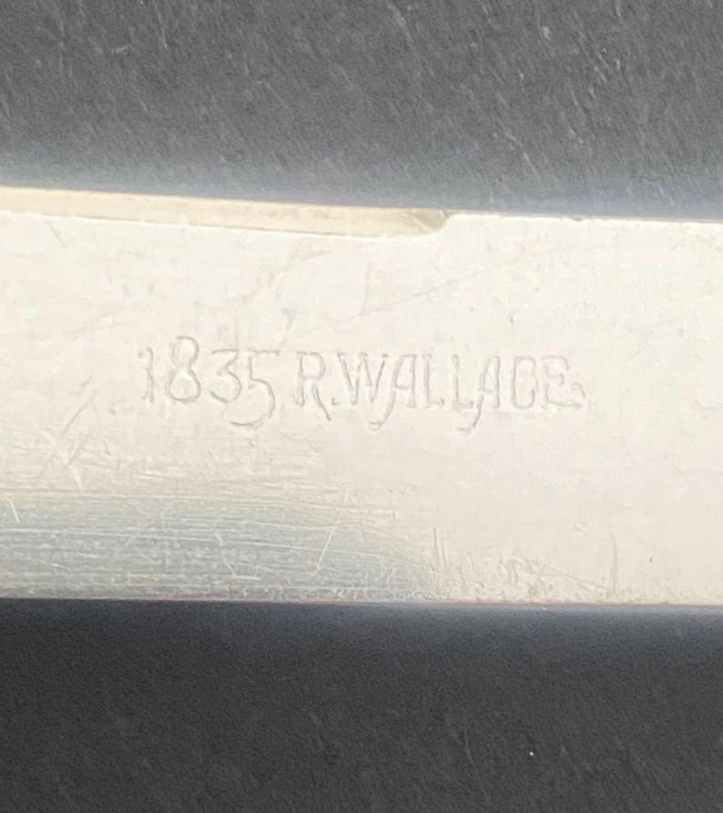 May include: Close-up of a silver metal object with the engraved text "1835 R. WALLACE". The metal surface has visible scratches and wear, indicating age. The text is in a serif font.