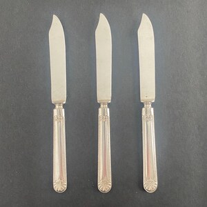 May include: Three silver butter knives with curved blades and ornate handles. The knives are arranged vertically against a dark background. The handles have a detailed, symmetrical design.