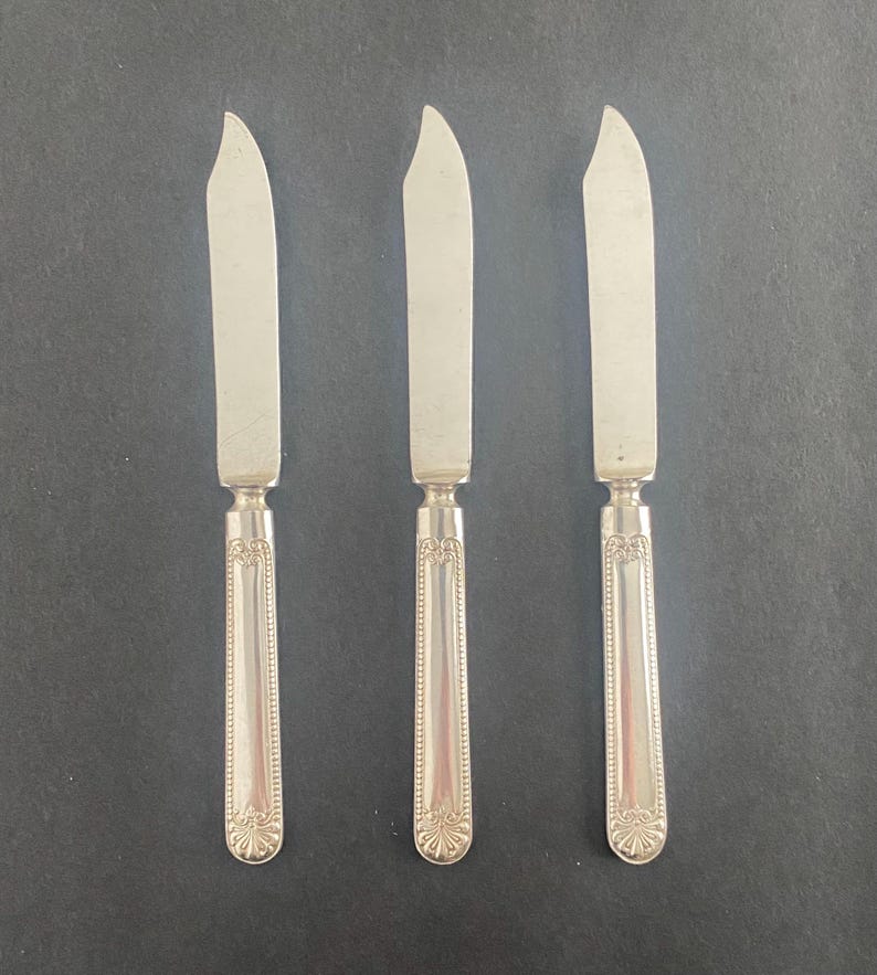May include: Three silver butter knives with curved blades and ornate handles. The knives are arranged vertically against a dark background. The handles have a detailed, symmetrical design.