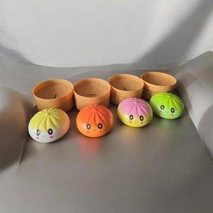 Peut inclure: Quatre petits pains jouets ronds et colorés avec des visages souriants et des joues roses, chacun présentant un dégradé de couleur distinct incluant jaune, orange, rose et vert, disposés devant quatre petits contenants circulaires marron clair.
