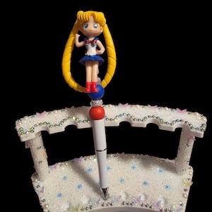 May include: A white pen holder with a decorative design, featuring a pen and a Sailor Moon figurine. The holder is embellished with rhinestones, pearls, and butterfly accents. The pen has a red top and a white barrel.