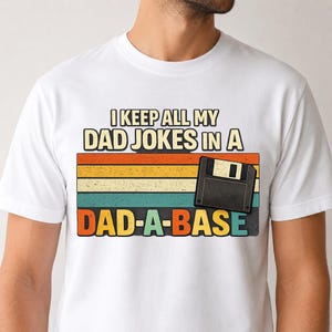 May include: White t-shirt with a retro design featuring the text "I KEEP ALL MY DAD JOKES IN A DAD-A-BASE". The design includes a floppy disk graphic and a colorful striped background.