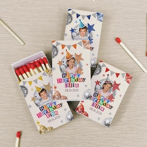 May include: Three rectangular matchboxes with white covers featuring a photo of a child, a party hat, disco balls, and colorful bunting. The text reads Happy Birthday Finn or Ella with the date 08.26.2020. One box is open, revealing red-tipped matches.