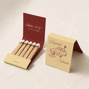 May include: Two matchbooks with cream and deep red covers. One open matchbook reveals wooden matches and text reading Malibu, CA JULY 24, 2027. The closed matchbook features a line drawing of a car with the names Benjamin and Amelia printed.