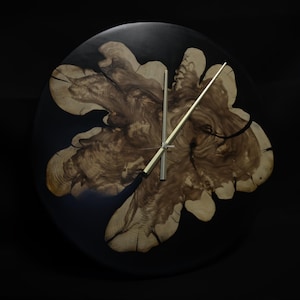 May include: A circular wall clock with a dark backdrop. The clock face showcases a natural wood design in shades of brown and tan, set in a black resin base. The clock hands are gold-coloured.