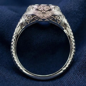 Könnte beinhalten: Nahaufnahme eines silbernen Rings mit aufwendigen Filigranarbeiten. Der Ring hat ein verziertes Design um das Band und eine dekorative Fassung. Der Ring ist vor einem dunkelblauen Hintergrund platziert, der das Glänzen des Silbers hervorhebt.