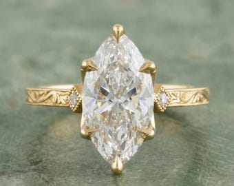 May include: A marquise-cut diamond engagement ring with a gold band. The ring features a large, clear diamond set in a four-prong setting. The band has an engraved floral pattern and small accent diamonds on each side of the main stone.