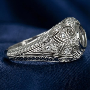 Puede incluir: Un anillo de plata ornamentado con un diseño de filigrana detallado. El anillo presenta un engaste central, probablemente para una piedra preciosa, rodeado de pequeñas piedras de acento brillantes. La banda y el engaste están intrincadamente tallados con patrones geométricos y florales.