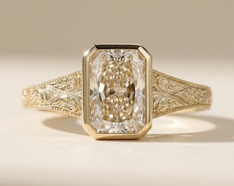 Pode incluir: Um anel de ouro com uma grande pedra preciosa retangular de cor clara. A pedra preciosa é engastada em uma moldura. A banda tem um design gravado e ornamentado. O anel é exibido em um fundo neutro.