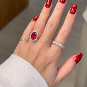 May include: A hand adorned with three rings: a plain gold band, a diamond eternity ring, and a gold ring featuring a red oval gemstone encircled by diamonds. The nails are painted a glossy red.
