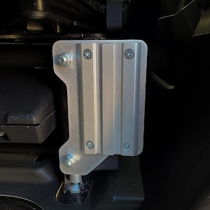 May include: A silver-colored metal mounting bracket with a vertical grooved channel design. The bracket is secured with four silver screws and two bolts with nuts, featuring a flat plate base that attaches to a dark, recessed mechanical structure.