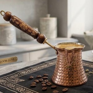 Könnte beinhalten: Eine Kupfer-Kaffeekanne, oder Cezve, mit einem Holzgriff mit floralen Schnitzereien. Die Kanne ist mit Kaffee gefüllt und steht auf einem dunklen Tuch mit verstreuten Kaffeebohnen. Der Griff ist etwa 15 cm lang.