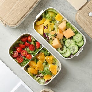 May include: Two white rectangular lunch boxes with wood-effect lids, filled with fresh salads and fruits. One box contains tomatoes, oranges, and lettuce. The other has mango, melon, and cucumber. A beige container with utensils is also visible.