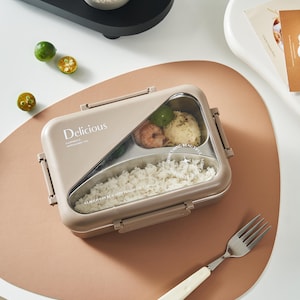May include: A beige rectangular lunch box with a clear lid, containing rice and other food items. The lunch box has the word "Delicious" printed on it. A white-handled fork rests on a pink mat next to the lunch box. A small bowl of fruit sits on a black tray.
