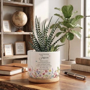 May include: A white ceramic planter with a succulent plant, featuring a floral design and the text "Good Morning Jessica". The planter sits on a wooden saucer. Other items include a globe, books, and framed art.
