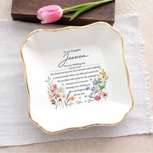 May include: A white ceramic dish with a gold rim, featuring floral illustrations and a heartfelt message. The text reads "To our Daughter Jessica" with a wedding date. A pink tulip rests nearby.