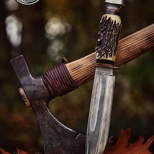 May include: A close-up of a hunting knife with a textured antler handle and a silver blade, resting on a wooden surface next to a small axe. The axe has a wooden handle and a dark metal head. The image has a blurred background.