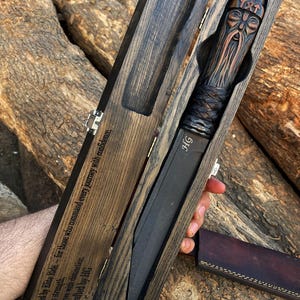 Könnte beinhalten: Ein schwarzer Dolch mit einem geschnitzten Holzgriff mit einem Gesichtsdesign, präsentiert in einer dunklen Holzkiste. Die Kiste hat eingravierten Text und wird offen gehalten. Der Dolch hat die Initialen "HG" auf der Klinge eingraviert.