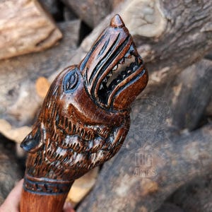 Puede incluir: Un bastón de madera tallado a mano con un diseño de cabeza de lobo. La cabeza del lobo está intrincadamente detallada, con las mandíbulas abiertas y los dientes visibles. La madera tiene un color marrón oscuro intenso y la talla es muy detallada.