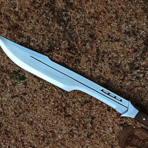 May include: A large, silver-colored blade with a sharp edge and a dark wooden handle. The blade has a distinctive shape with a pointed tip and a series of cutouts along the spine. The handle is made of dark wood and is secured with rivets.