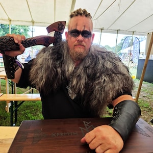 Puede incluir: Una persona con atuendo vikingo, sosteniendo un hacha de madera tallada y usando un cuello de piel y brazaletes de cuero. Una caja de madera con diseños grabados y el texto "XFRMTN" está frente a ellos. La persona tiene pintura facial.