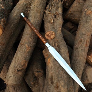May include: A long, bladed spear with a polished steel blade and a wooden handle. The handle is dark brown with small, evenly spaced silver accents. A small, round wooden emblem with text is also present.