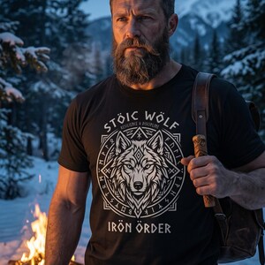 以下が含まれることがあります： オオカミのグラフィックと「STOIC WOLF RESILIENCE & DISCIPLINE IRON ORDER」の文字が入った黒いTシャツ。 デザインは白で、円形のフレームの中にオオカミの頭が描かれています。 その人は茶色の革のバックパックを持っています。
