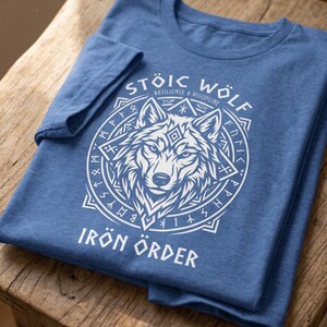 以下が含まれることがあります： 白いオオカミのグラフィックが円形デザインで描かれた青いTシャツ。「STOIC WOLF」と「IRON ORDER」の文字がオオカミのイラストの上と下にプリントされています。Tシャツは木製の表面に折りたたまれています。