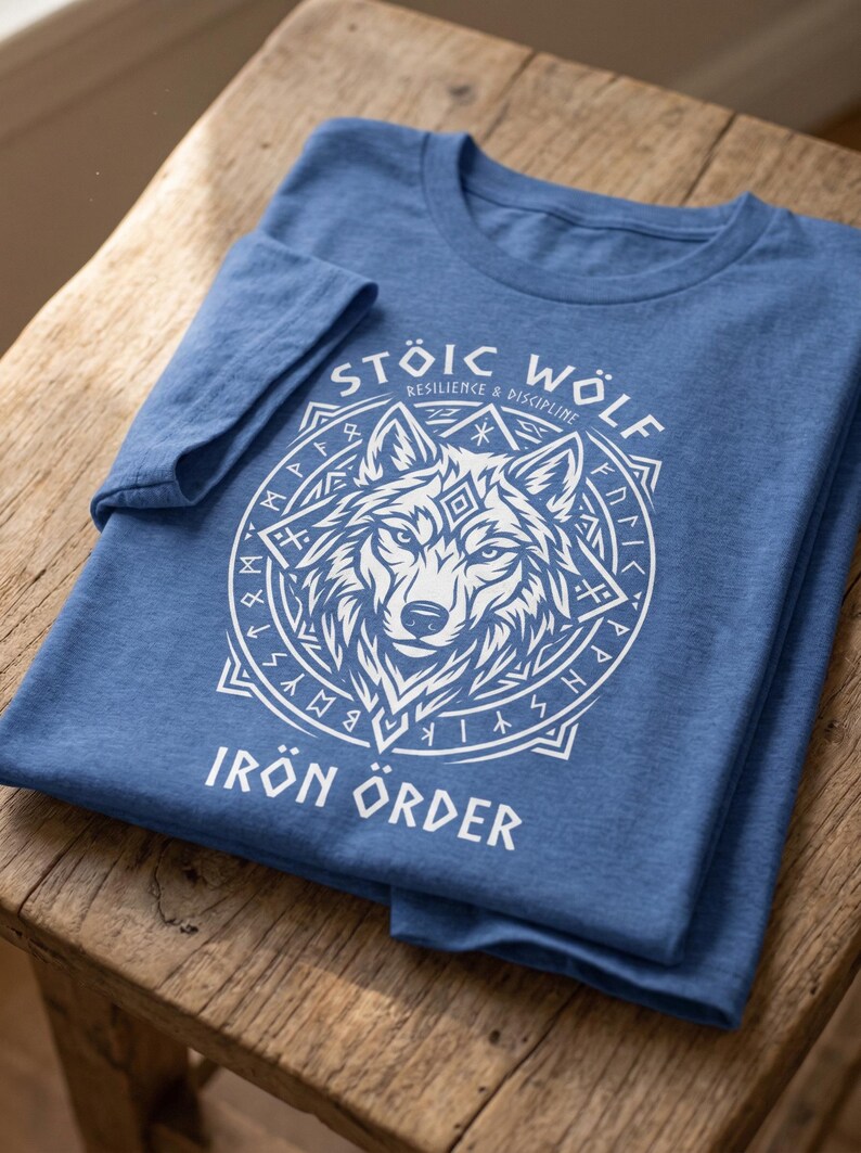 以下が含まれることがあります： 白いオオカミのグラフィックが円形デザインで描かれた青いTシャツ。「STOIC WOLF」と「IRON ORDER」の文字がオオカミのイラストの上と下にプリントされています。Tシャツは木製の表面に折りたたまれています。