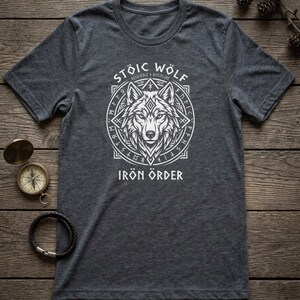 以下が含まれることがあります： 白オオカミのデザインと「STOIC WOLF IRON ORDER」の文字が入った濃い灰色のTシャツ。デザインは、ルーン文字が入った円形のフレームの中のオオカミの頭部が特徴です。コンパスとブレスレットが木製の表面にあります。