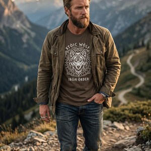 以下が含まれることがあります： オオカミのグラフィックと「Stoic Wolf Iron Order」の文字が入った茶色のTシャツを着た男性。彼はまた、緑色のジャケットとブルージーンズを着用しています。背景には山の風景が広がっています。
