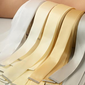 May include: A selection of satin ribbons in various shades of white, cream, and gold. Some ribbons have a decorative rhinestone trim. The ribbons are rolled and fanned out, showcasing their smooth texture and color variations.