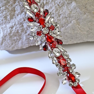May include: A red and silver rhinestone bridal belt with a satin ribbon. The belt features a floral design with red and clear rhinestones of various shapes and sizes. The ribbon is a bright red color.
