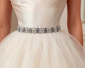 Op de afbeelding: Een crèmekleurige jurk met satijnen lijfje en een brede, decoratieve riem in de taille. De riem is versierd met een herhalend patroon van heldere kristallen, kleine witte parels en blauwe marquise-geslepen stenen in een zilverkleurig montuur.