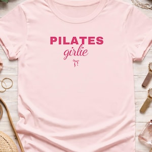 Può includere: T-shirt rosa chiaro con la scritta "PILATES girlie" in rosa più scuro. La parola "PILATES" è in grassetto, senza grazie, mentre "girlie" è in corsivo con un piccolo fiocco. La maglietta ha maniche corte e scollo tondo.