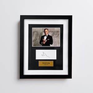 May include: Framed photograph of Daniel Craig in a tuxedo, with a signature and nameplate. The black frame surrounds a white matting, with a photo of Craig in a black tuxedo. Below the photo is a white card with a signature and a gold nameplate.