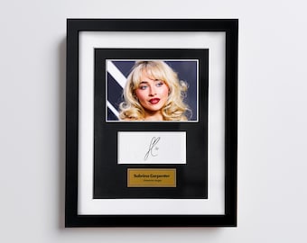 May include: Framed photograph of Sabrina Carpenter with a signature and a gold plaque that reads "Sabrina Carpenter, American singer". The frame is black with a white matting.